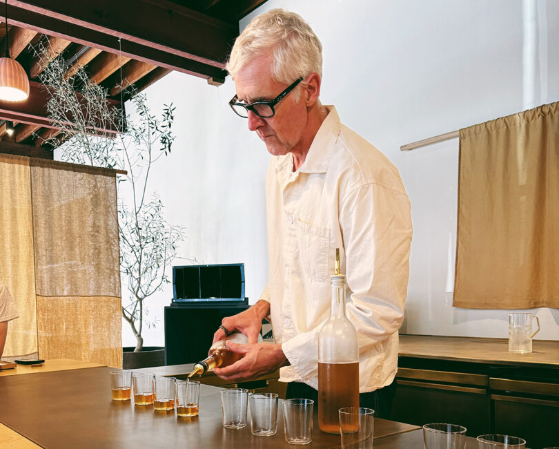 Blue Bottle Coffee’s founder, James Freeman, pouring coffee leaf steeped tea into ten glasses behind the Blue Bottle Studio bar. He's wearing a white button up long sleeve shirt and dark eyeglasses.