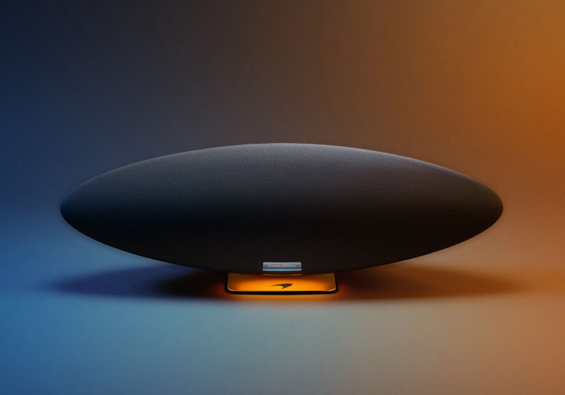 Front facing wide shot of Bowers & Wilkins Zeppelin McLaren Edition wireless speaker, finished in Galvanic Grey body and Papaya Orange, set against a blue to orange illuminated gradient lighting.