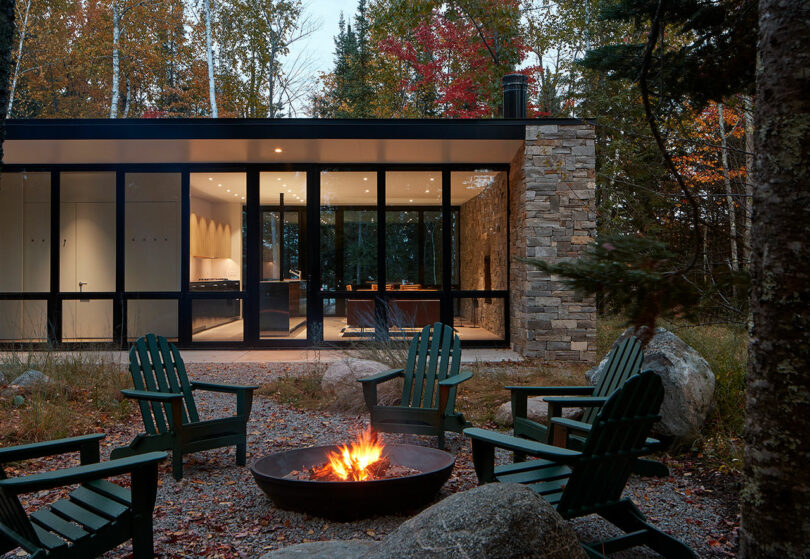 close up exterior shot of a one-story rectangular stone house surrounded by forest with fire going in pit