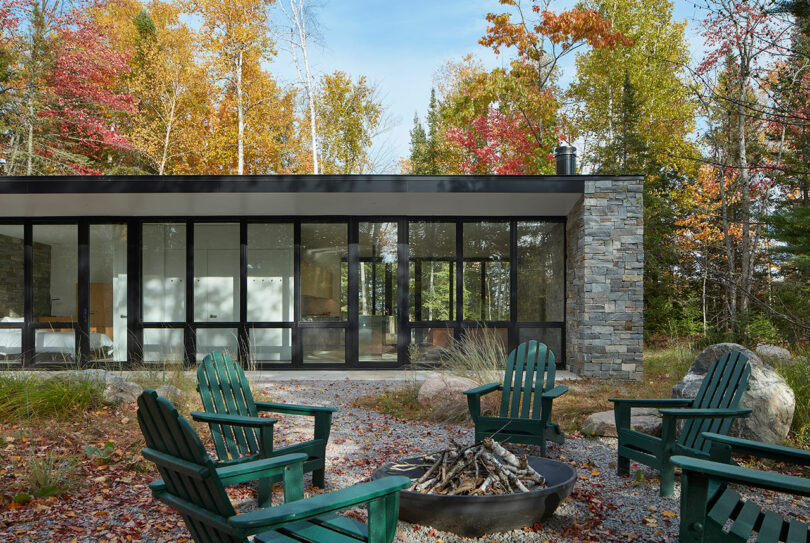 closeup exterior shot of a one-story rectangular stone house surrounded by forest with green chairs surrounding fire pit
