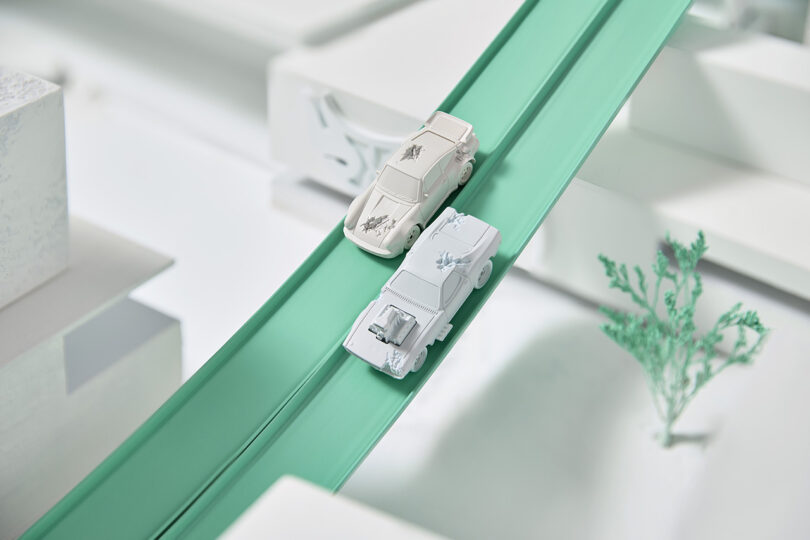 Stone white Daniel Arsham x Hot Wheels collection Porsche and Dodge Charger models made from Silkstone racing side by side down a winding downward across a stretch of green plastic toy track.