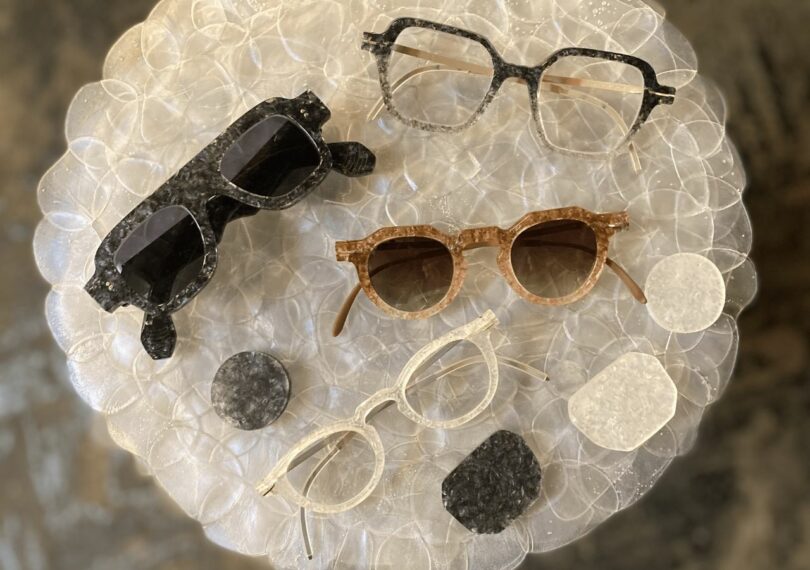 Two pairs of glasses and two pairs of sunglasses and laid next to material samples on a surface that appears to be made from overlapping opaque disks.