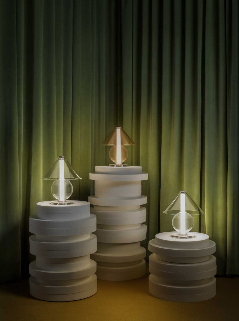 three all glass lamps with round base and cone-shaped shade illuminated on pedestals in front of an olive green backdrop