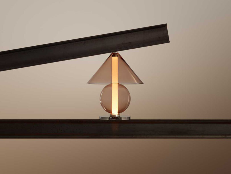 all glass lamp with round base and cone-shaped shade illuminated balanced between two pieces of wood