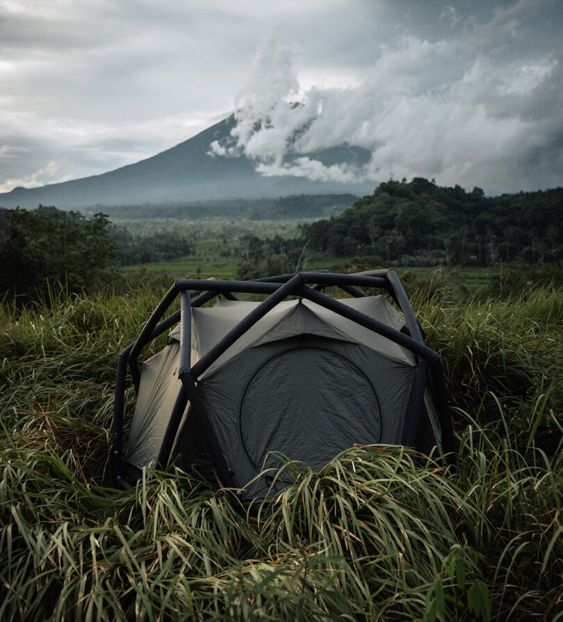 Geodesic dome shaped tent pitched out in tropical grass covered landscape with large cloud covered mountains in the background