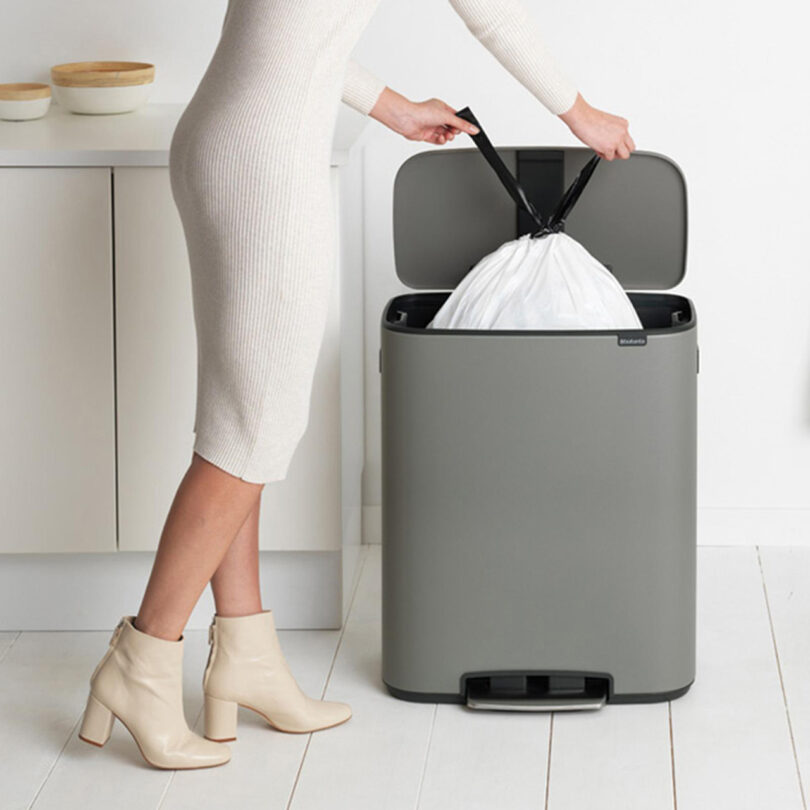 woman in form fitting white dress and boots reaching into large gray trash bin stationed in a white modern kitchen, pulling out large white plastic garbage bag 