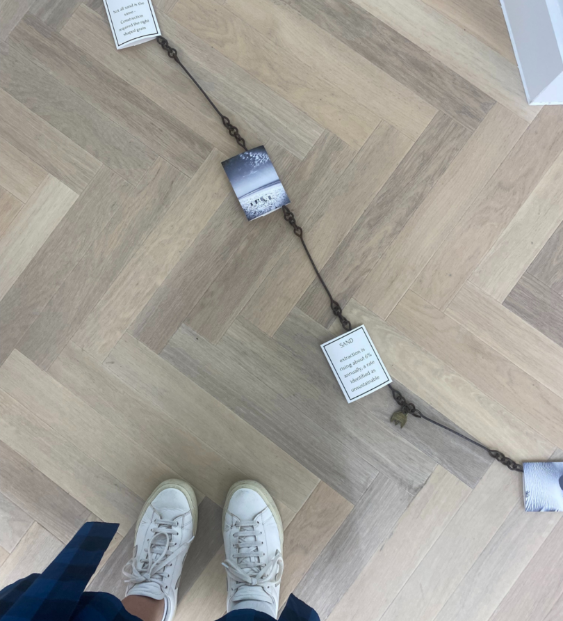 A chain with pieces of card containing text and images attached to it laid on the floor. Two feet in white trainers are just visible. 