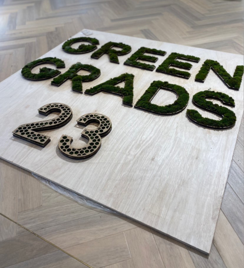 The words "Green Grads 23" are made out with moss or another form of growing matter on a wooden board on the floor.