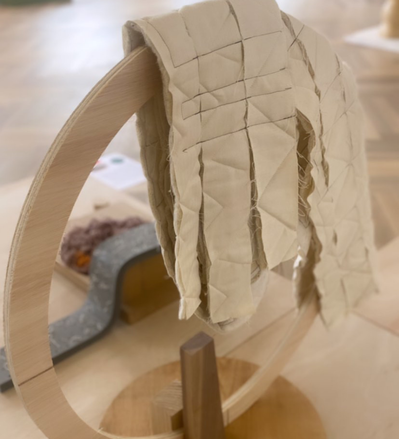 A vertical hoop of plywood with offcuts of quilted fabric draped over it. 