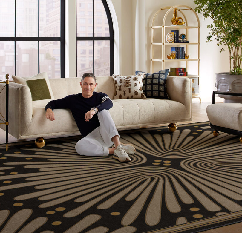 Designer Jonathan Adler seated upon the floor rug leaning against a contemporary sofa with a large arched window and bookcase in the background.
