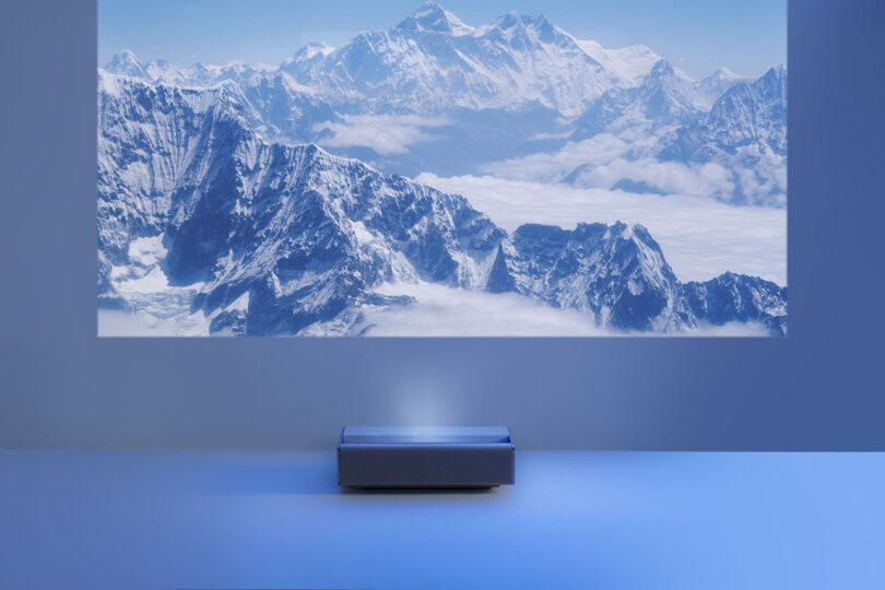Front view of the soft earth tone fabric covered XGIMI Mira ultra short throw projector. Unit has soft curves and brushed metal edge detailing, and it is projecting an image of enormous snowcapped mountains positioned on the floor and in a darkened room.