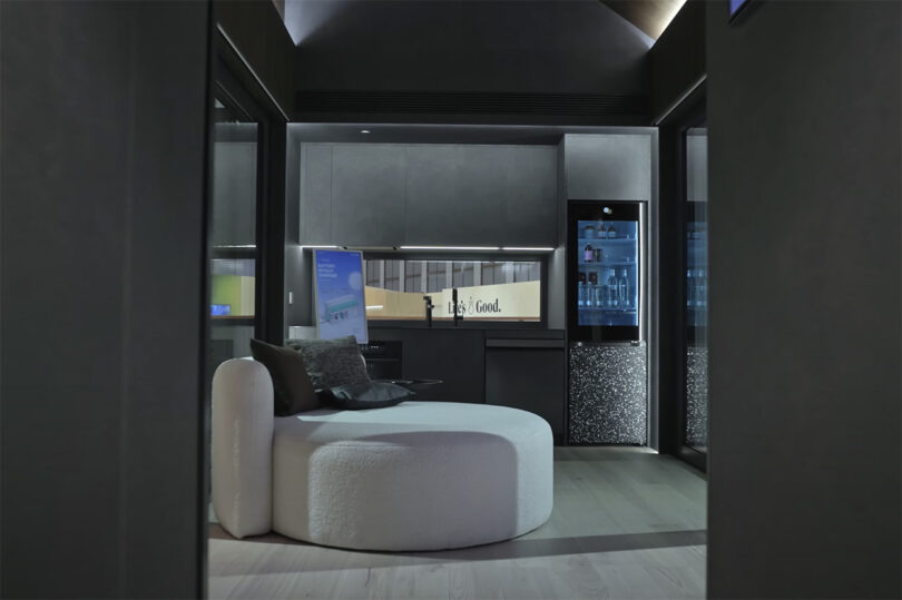 Dark interior of Exterior of LG Smart Cottage showcase home at IFA 2023 with LG television in vertical display mode and round chaise lounge with small integrated kitchen in the background.