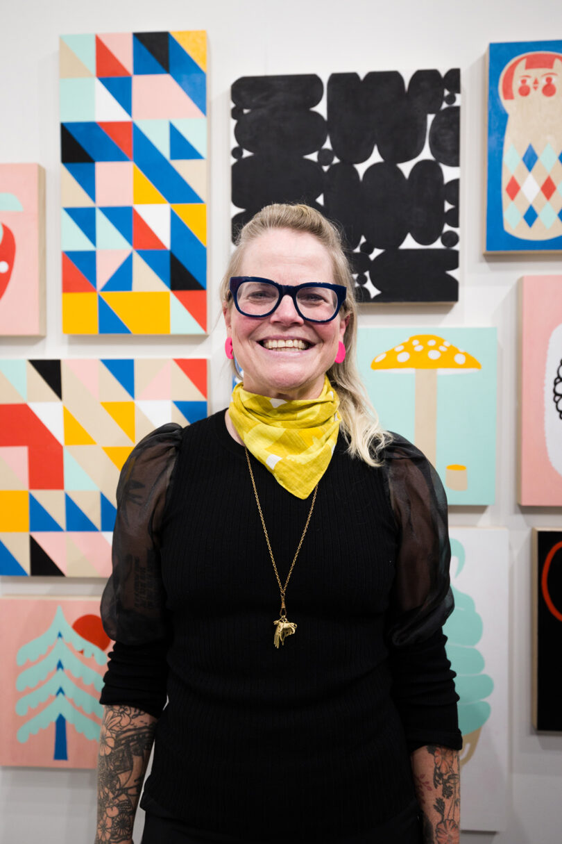 light-skinned woman with blonde hair and glasses stands in front of gallery wall covered in bright illustrative pieces of art