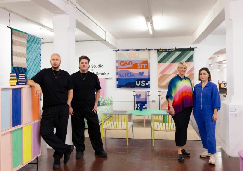 Two white men and two white women all in their 30s/40s stand in front of a colorful array of furniture and wall hangings included a blanket that reads "You Can Sit With Us." 