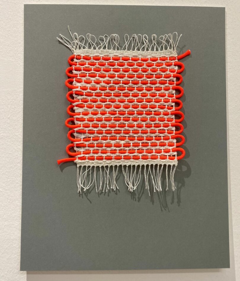 A bright orange cord is woven into an off-white yarn to create a striking wall hanging / fabric patch on a grey background. 