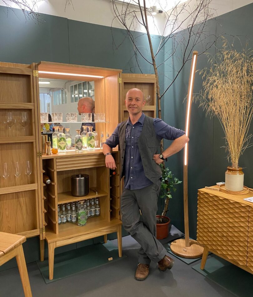 A white man in his 40s stands leaning on a wooden drinks cabinet. The branch of a tree is behind him and to his left a lower wooden side cabinet. 