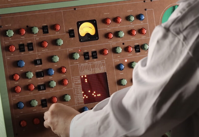 Man in white lab coat reaching to turn one of the dials of the ATOM-IC green and brown wood cabinet synthesizer with a small LED matrix display showing 14 dots of light to represent sources and inputs.