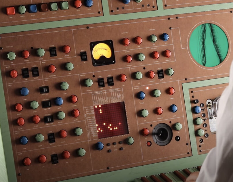Detail close up of green and brown wood cabinet synthesizer with small keyboard and layout of numerous colorful dials, switches, and buttons. Oscillator screen shows a wavy pattern.