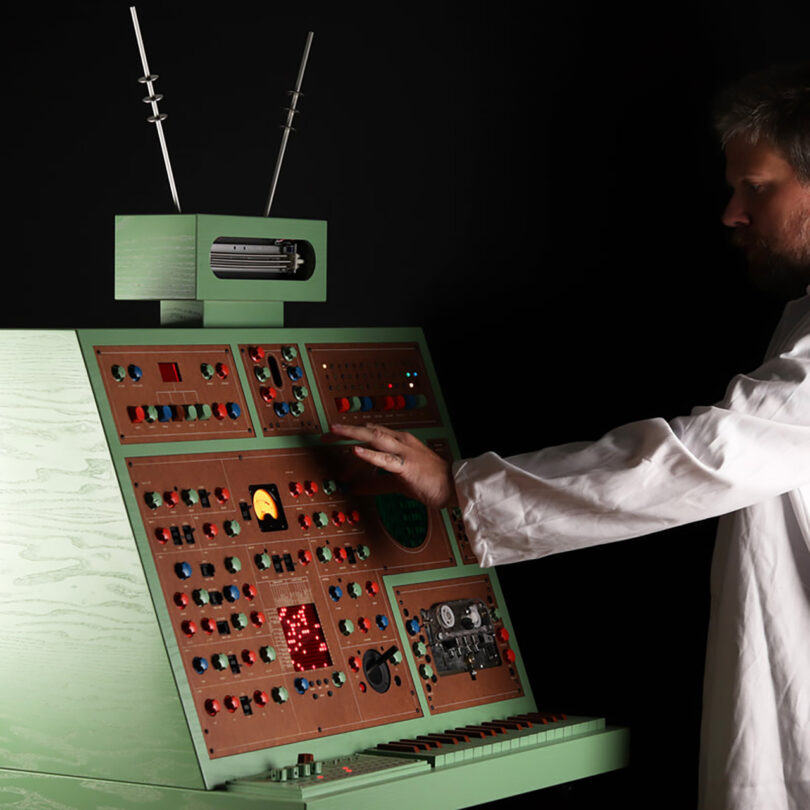 Green and brown wood cabinet synthesizer with small keyboard and layout of numerous colorful dials, switches, and buttons all topped with two metal telescoping antennas. The designer is in a white lab coat and reaching to press a button or turn a dial.