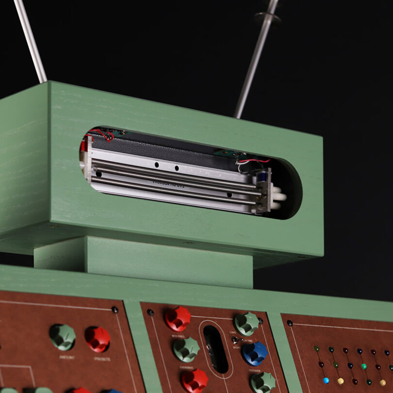 Detail of antenna topped ATOM-IC green and brown wood cabinet synthesizer with a section of the numerous red, green and blue dials, switches, sliders and buttons controls visible underneath.