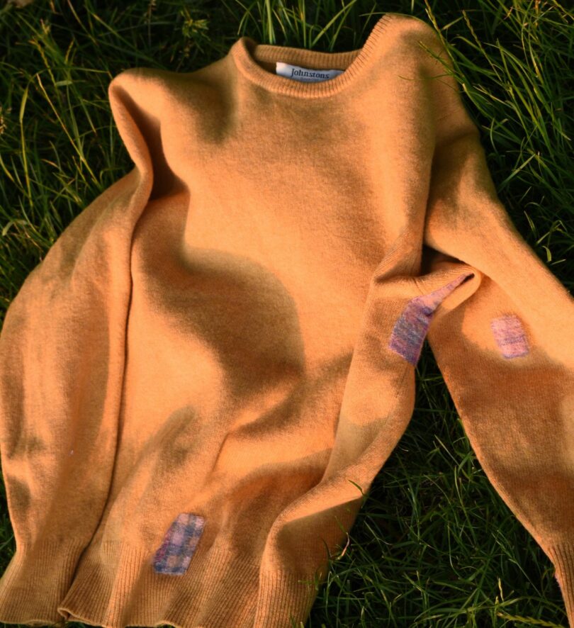 An orange knitted sweater lies on the grass – it has three pink and purple checked patches