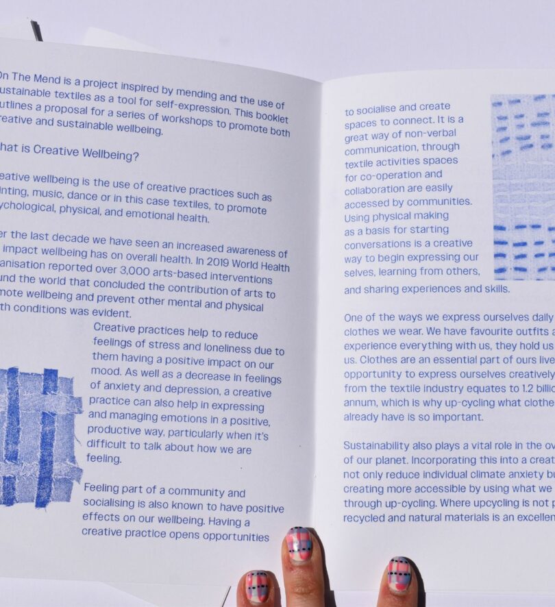 Three white fingers hold open a printed publication that includes words and images of mended fabric – one headline reads "what is creative wellbeing?"