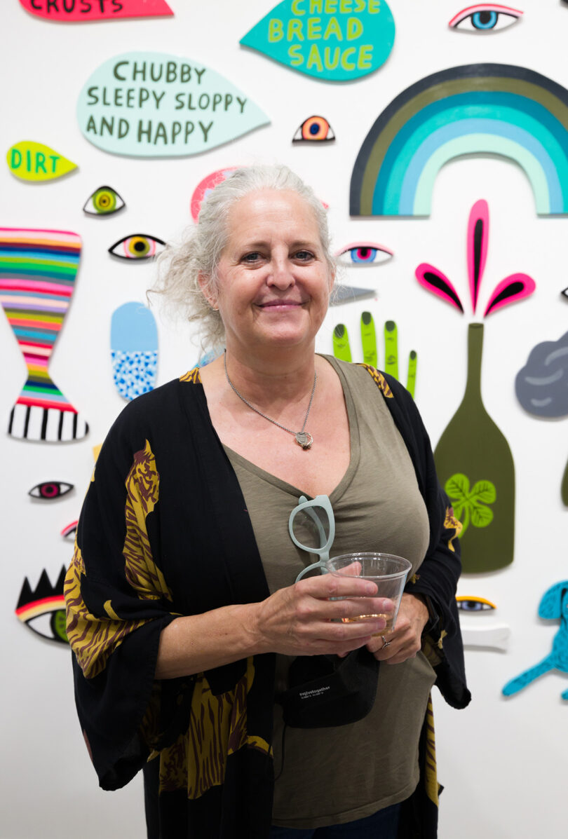 light-skinned woman with white hair pulled up stands in front of a gallery wall filled with small, colorful pieces of painted wood objects and speech balloons