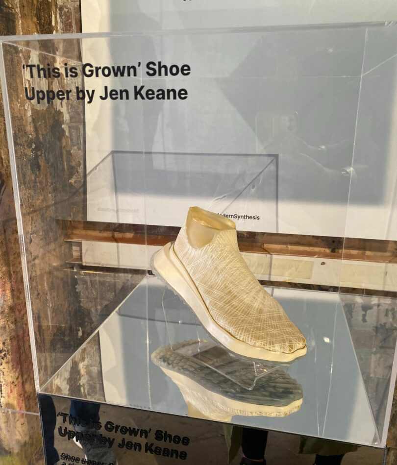 A white woven shoe in a glass box with the words "This is Grown" Shoe Upper by Jen Keane printed on the front.