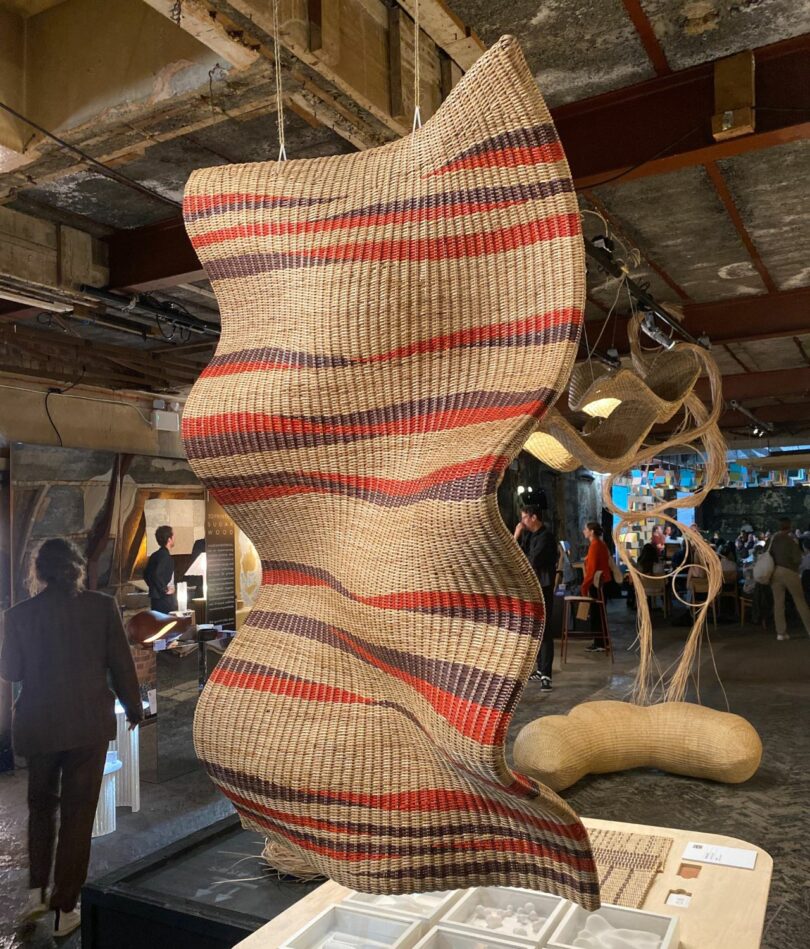 An undulating wicker form hangs from the ceiling. It is mostly beige-brown with red and black stripes coming in from either side. It is hanging in a warehouse roof space.