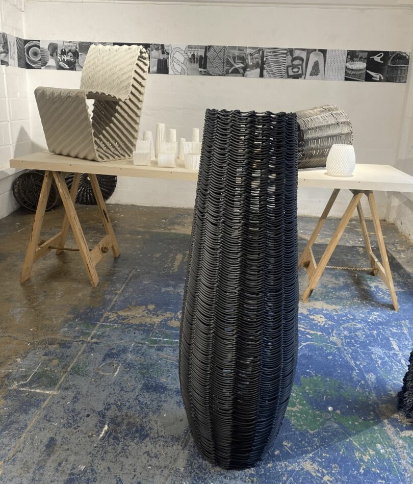 A tall black vessel stands in the foreground with a cream chair in a trestle table behind. Both are made from overlapping strings and loops of plastic. 
