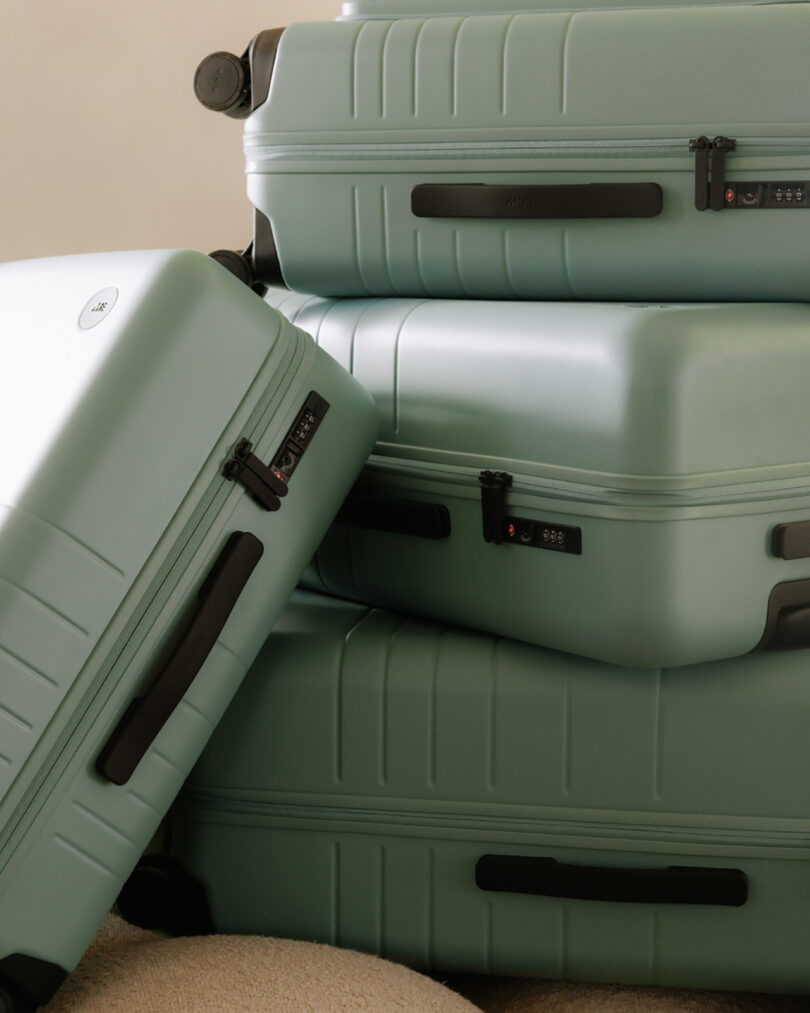 A pile of Monos carry-on and check-in hard case luggage in Sage Green, three stacked onto one another on their side, one leaning against the tother three vertically.
