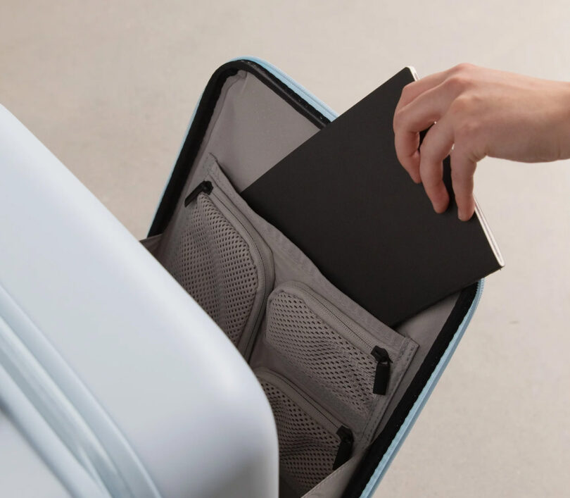 Person placing a black notebook into the front foldout compartment storage of the Monos Carry-On Pro in Blue Haze color finish, with the front case door ajar