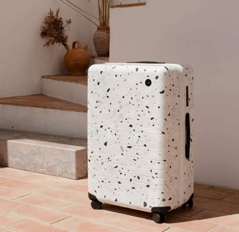 Monos check-in hard case luggage in a Terrazzo style design, stationed near a set of outdoor stairs with clay tiling and white walls.