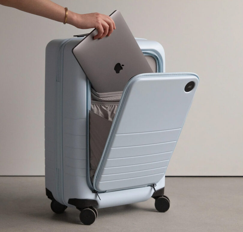 Woman placing an Apple MacBook Air into the front foldout compartment storage of the Monos Carry-On Pro in Blue Haze color finish, with the front case door ajar