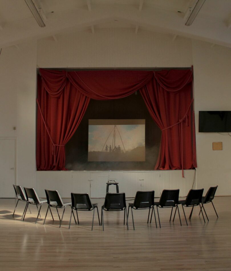 Nine plastic chairs are arranged in a arc in front a stage, which is surrounded by drawn red curtains and onto the backdrop of which is projected an image.