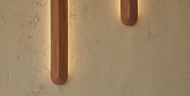 A simple, elongated wall light, offered in two lengths – 400mm and 900mm with a linear shield-like form characterized by a central sharp line or ‘pinch’ that allows light to fall differently on each side of the terracotta.