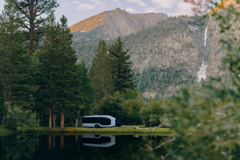 White Pebble Flow RV trailer unhitched and parked near a pond overlooked by large looming forest covered mountains.