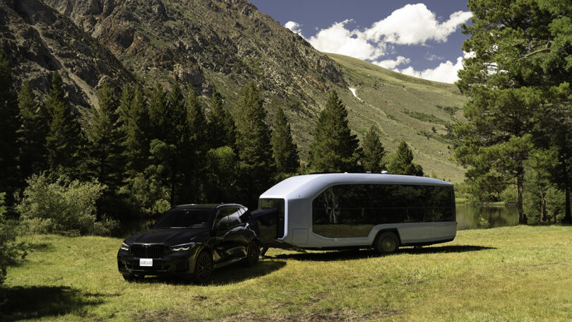 Black SUV with a white Pebble Flow RV trailer behind it parked in a grassy known within a mountain forest setting with blue skies and a few clouds overhead.