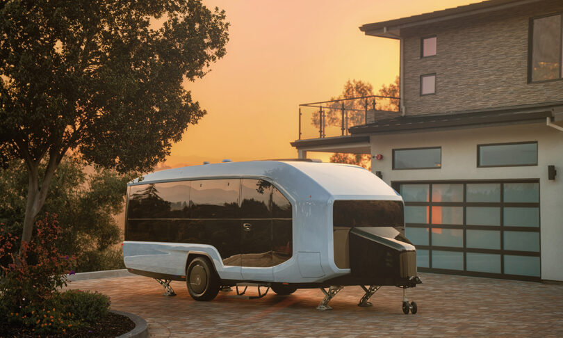 Detached white Pebble Flow RV trailer parked in front of large contemporary home with glass paned style garage door. Sunset skies loom in the background.