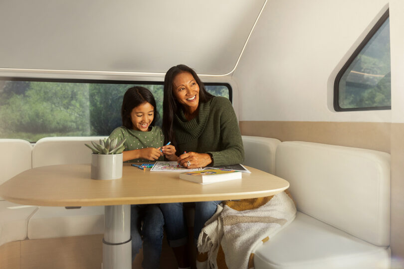 Woman and daughter seated at the back of the Pebble Flow RV trailer's dining and seating area, working on color books while both smiling.
