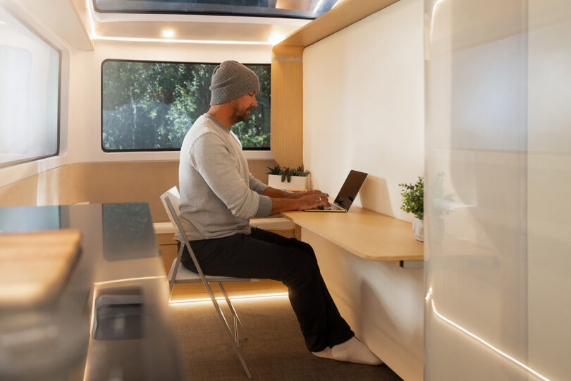 Man in gray sweater and beanie seated at a narrow floating desk working from his laptop inside the Pebble Flow RV.