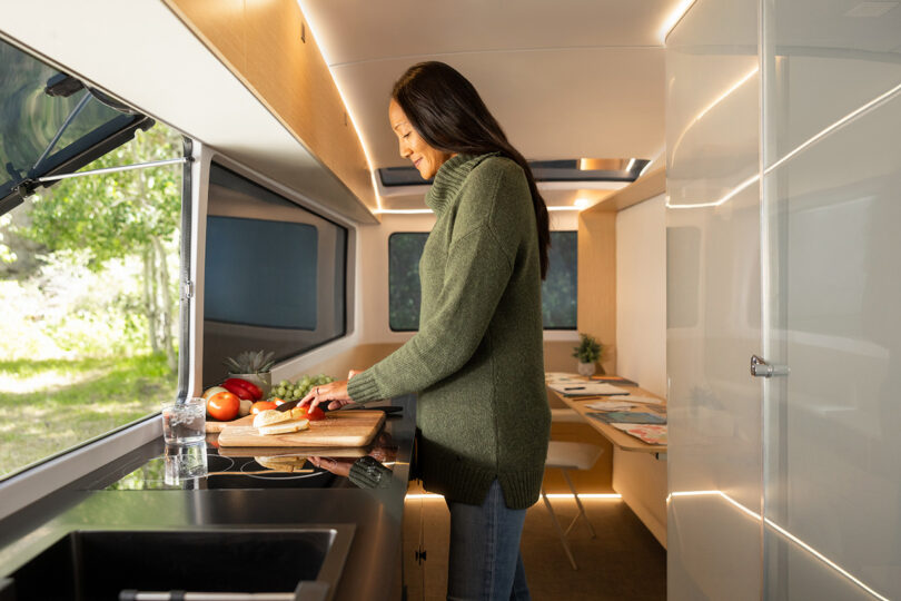Woman with long black hair and in green turtleneck sweater dicing vegetables on a cutting board inside the galley kitchen of the Pebble Flow RV trailer.