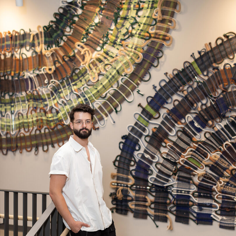 light-skinned man with dark hair and facial hairs leans against a railing in front of two large handwoven art pieces hanging on a white wall