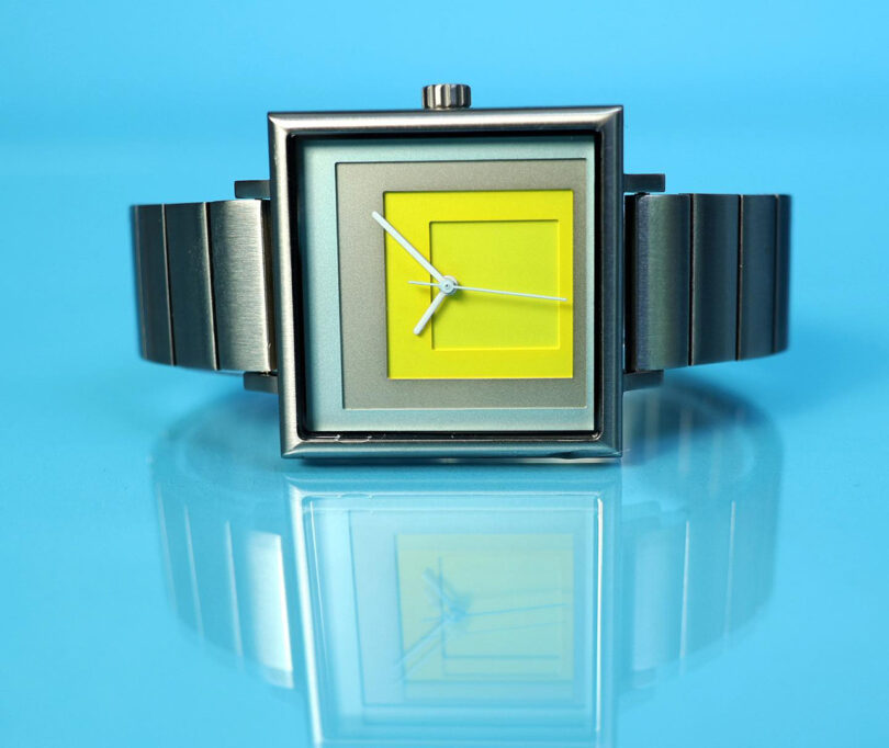 Four layers of yellow, blue, and steel laser cut squares of color inside a 35mm stainless steel watch case with stainless steel band inspired by the artwork of Josef Albers. The watch is laying on its side with crown facing upward, across a shiny light blue surface.