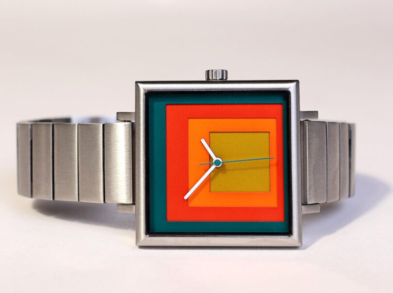 Four layers of red, orange, and green laser cut squares of color inside a 35mm stainless steel watch case with stainless steel band inspired by the artwork of Josef Albers. The watch is laying on its side with crown facing upward, across a white surface.