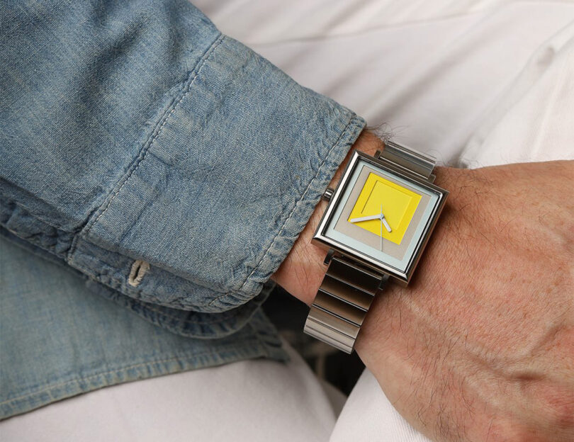 Close up of four layers of yellow, blue, and steel laser cut squares of color inside a 35mm stainless steel watch case with stainless steel band inspired by the artwork of Josef Albers on man's wrist; man is wearing denim long sleeve shirt with cuff unbutton and white pants.