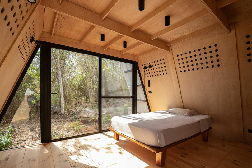 vista interior da cabana moderna com paredes de madeira inclinadas e cama minimalista em frente às janelas
