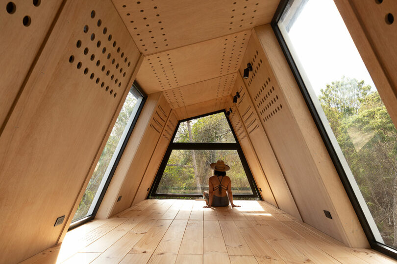 interior de uma cabana minimalista com paredes de madeira com uma mulher sentada de costas olhando para as árvores