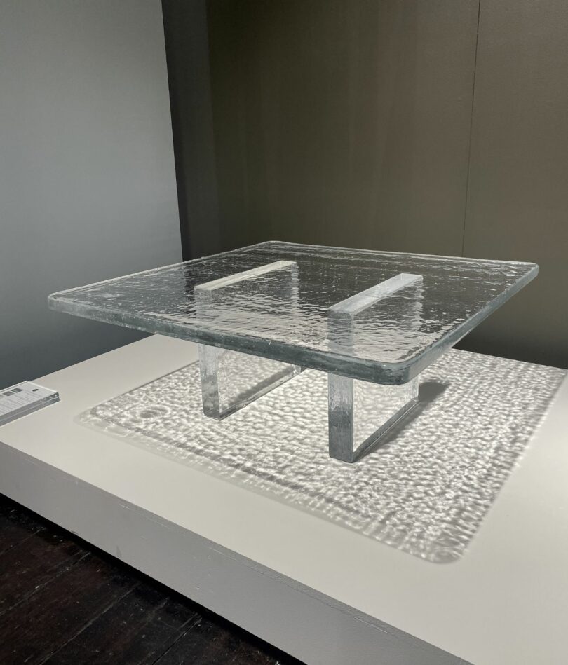 A table is made from three solid slabs of glass, two on end as legs and one laid across them as a top. Dappled light is cast through the table onto a white plinth beneath.