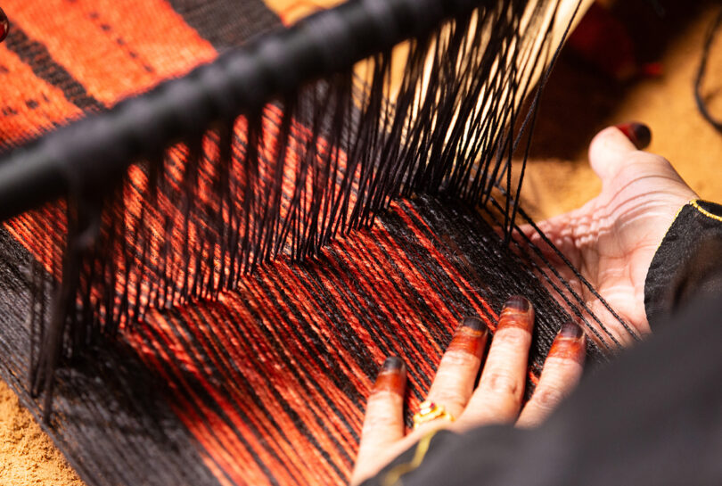 light-skinned hand working on a loom strung with black and rust colored yarns